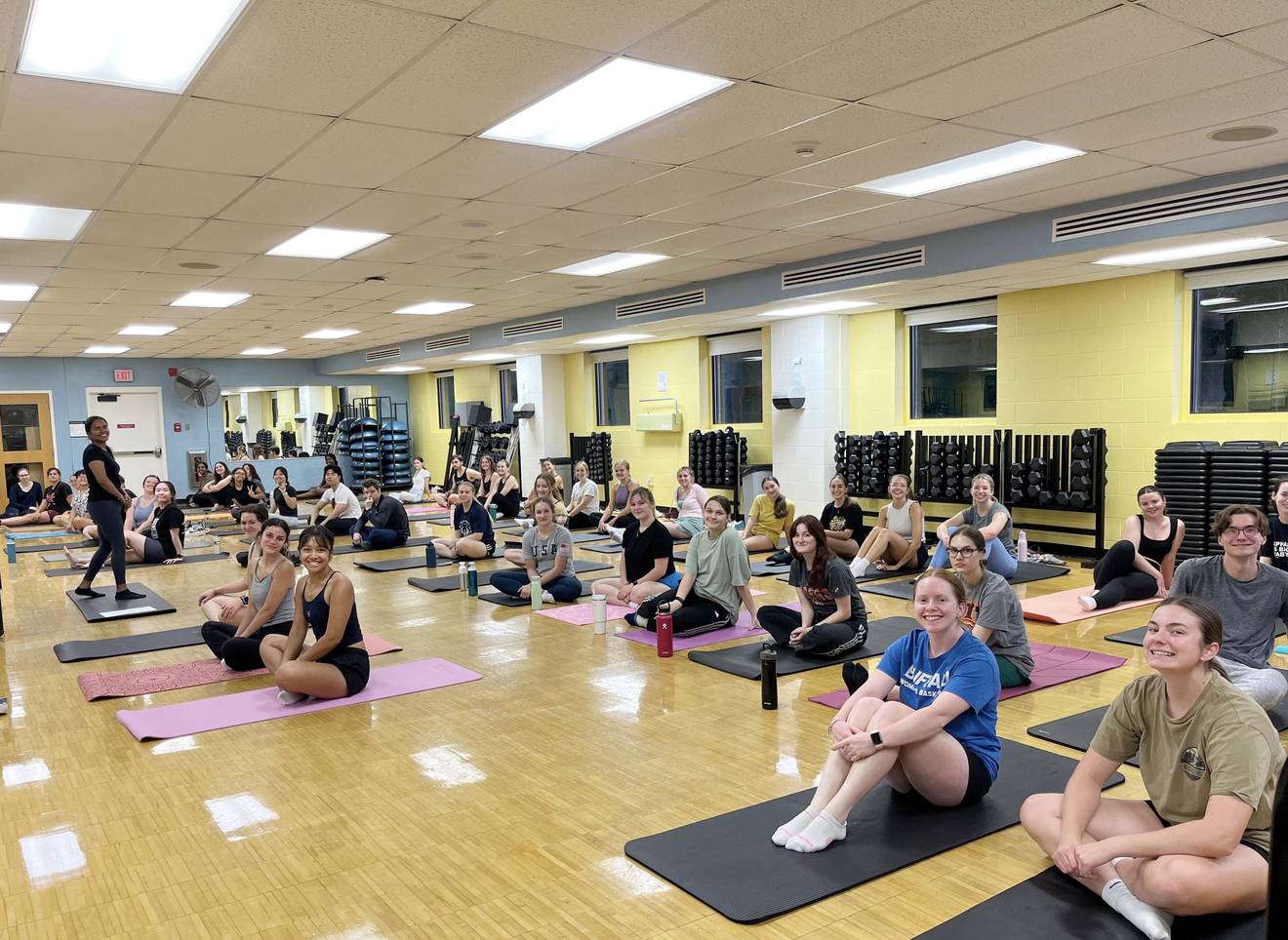 A full studio of students seated on mats before a yoga class begins